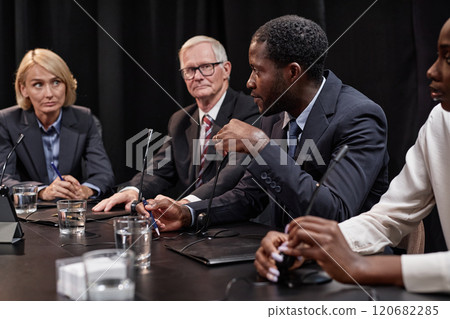 Side view of confident African American politician speaking into microphone debating with female colleague about initiative while sitting in line with other officials at meeting table, copy space Side view of confident African American politician speaking into microphone debating with female colleague about initiative while sitting in line with other officials at meeting table, copy space 120682285