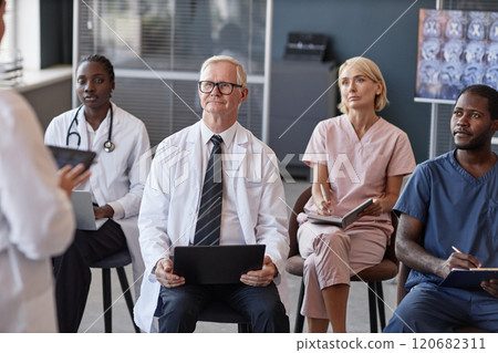 Senior male head physician listening to colleagues presentation with diverse team of doctors while participating in seminar at conference room 120682311