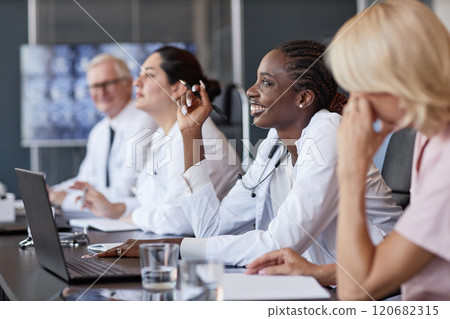 Side view of positive African American female clinician asking colleague follow up question after presentation in group seminar while sitting at table in medical office, copy space 120682315