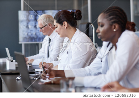 Side view of Middle Eastern female clinician listening to colleagues presentation keeping writing records while sitting at meeting table in medical office, copy space 120682316