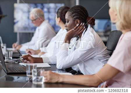 Side view of African American female clinician listening to colleagues presentation keeping records of working meeting while sitting at table in medical office, copy space 120682318