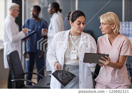 Medium shot of female Middle Eastern surgeon in conversation with colleague consulting medical records while looking at tablet computer screen following group seminar at office, copy space 120682321