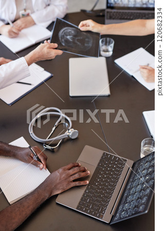 Vertical shot of Black man writing notes on paper keeping fingers on laptop touchpad checking X ray scan imaging on screen while sitting at meeting table with colleagues at medical office, copy space 120682334