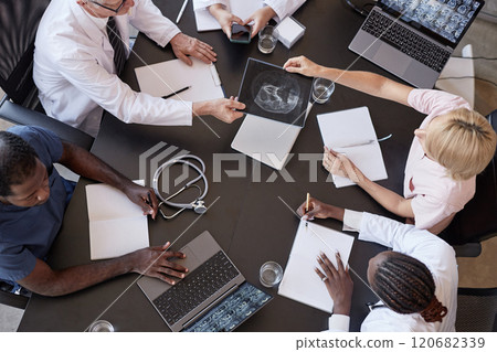 Top view of woman passing CT image of patients skull to chief physician discussing X ray examination results with colleagues, while sitting at table with laptops during meeting at medical office 120682339