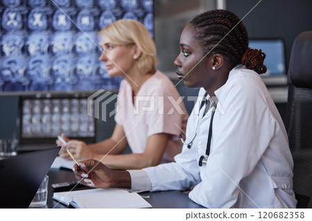 Side view of African American female neurologist in group discussion at seminar listening to colleagues opinions while writing notes in notebook sitting at meeting table at medical office, copy space Side view of African American female neurologist in group discussion at seminar listening to colleagues opinions while writing notes in notebook sitting at meeting table at medical office, copy space 120682358