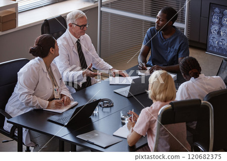 High angle shot of diverse group of professional clinicians thoroughly studying anamnesis of patients disease sitting at meeting table, while collaboratively planning treatment at medical office High angle shot of diverse group of professional clinicians thoroughly studying anamnesis of patients disease sitting at meeting table, while collaboratively planning treatment at medical office 120682375