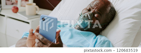 Elderly man wearing oxygen mask and using smartphone on hospital bed while looking at screen, bedside table with apple, mug, and other items in background Elderly man wearing oxygen mask and using smartphone on hospital bed while looking at screen, bedside table with apple, mug, and other items in background 120683519