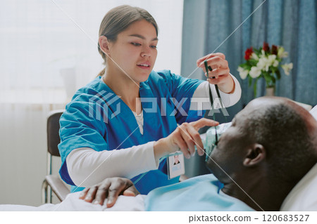 Nurse attending to senior patient by checking his condition using diagnostic device, hospital room background with medical equipment and floral decorations visible Nurse attending to senior patient by checking his condition using diagnostic device, hospital room background with medical equipment and floral decorations visible 120683527