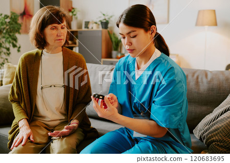 Nurse sitting on sofa while measuring elderly woman's blood pressure with digital device in cozy living room setting with plants and soft lighting Nurse sitting on sofa while measuring elderly woman's blood pressure with digital device in cozy living room setting with plants and soft lighting 120683695