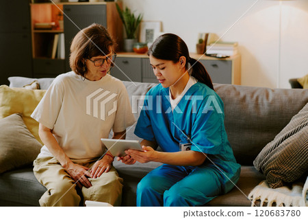 Nurse sitting on couch with elderly woman, discussing something on digital tablet in living room setting Nurse sitting on couch with elderly woman, discussing something on digital tablet in living room setting 120683780