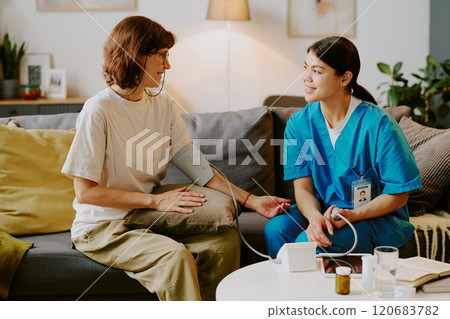 Elderly person having health checkup from smiling nurse in living room, nurse wearing scrubs measuring blood pressure while engaging in conversation 120683782