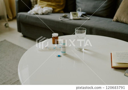 Various medication bottles, glass of water and open book arranged on white table in cozy living room with gray couch in the background 120683791