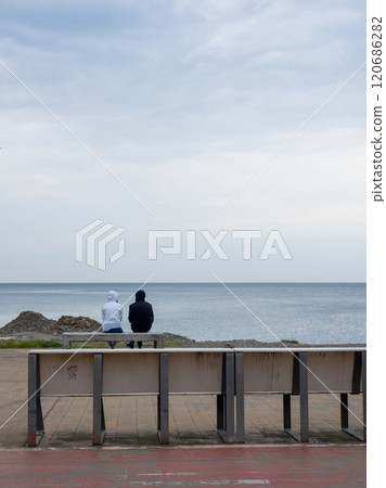 A couple is sitting on a bench by the sea. Cloudy weather at the resort. Not the season for tourism. Recreation of people on the coast. 120686282