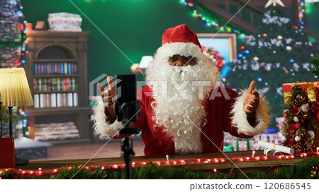 Santa Claus live streaming video blog sharing Christmas traditions from his festive North Pole cabin. Saint Nick in his red suit engages with online followers during the holiday season. Camera A. 120686545
