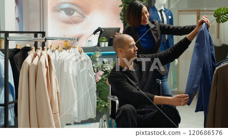 Asian friend assisting middle eastern wheelchair user in buying new clothes, looking at the new fashion collection on display at the clothing store. Man with disability shopping casual wear. Camera A. Asian friend assisting middle eastern wheelchair user in buying new clothes, looking at the new fashion collection on display at the clothing store. Man with disability shopping casual wear. Camera A. 120686576