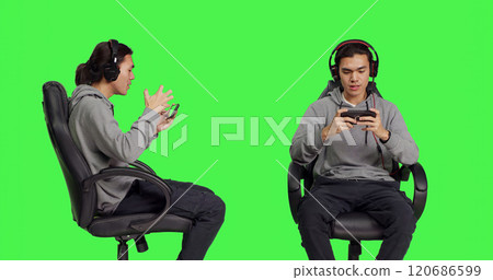 Asian gamer engaging in rpg contest online using mobile phone gameplay, having fun with other players on internet server. Young man enjoying gaming competition against greenscreen backdrop. 120686599
