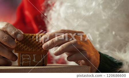 Father Christmas decorating gingerbread house and cookies with colorful icing in North Pole cabin. Santa enjoying the holiday tradition with festive activity, handmade desserts. Close up. Camera B. 120686600