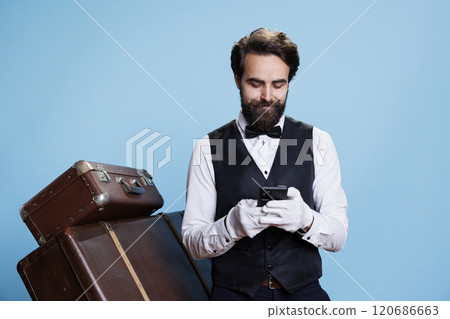 Hotel staff poses in studio with trolley baggage while he is cruising through social media and messaging. Doorkeeper or bellhop in official attire using phone app for surfing web pages. 120686663
