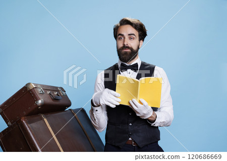 Wise bellhop with tie reads novel story, having fun with his hobby while he works as hotel porter. Doorman in suit reading literature, fiction or poetry for knowledge, guarding pile of suitcases. Wise bellhop with tie reads novel story, having fun with his hobby while he works as hotel porter. Doorman in suit reading literature, fiction or poetry for knowledge, guarding pile of suitcases. 120686669