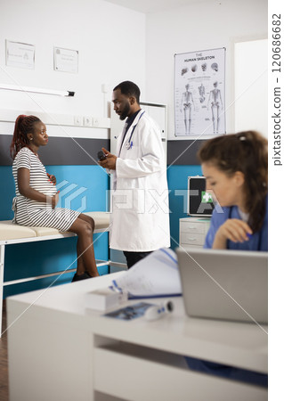 In background doctor with bottle of pills speaks to pregnant black woman while young female nurse reads medical records. Male physician provides prescribed supplements to expectant mother. 120686682
