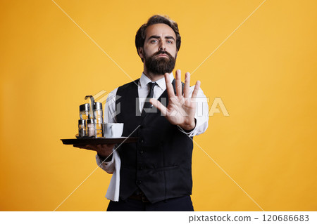 Skilled butler does stop sign with palm on camera, expressing disapproval and dismissing something. Young man with waiter occupation shows rejection and disagreement, not accept. 120686683