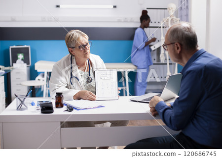 Senior doctor discussing treatment plans on tablet with elderly white man at clinic appointment. Retired female physician using digital device to clarify medical details for male pensioner patient. 120686745