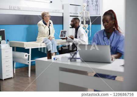 Black doctor uses tablet to explain healthcare options to old white woman focusing on treatment diagrams. Elderly patient and male physician carefully review medical details on digital device. 120686756
