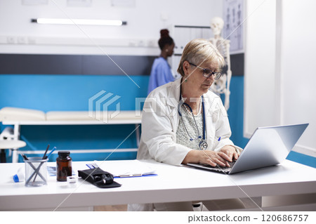Retired general practitioner sitting and using laptop in hospital room in preparation of patient appointments. Elderly female doctor checking emails and reviewing medical records in clinical office. 120686757