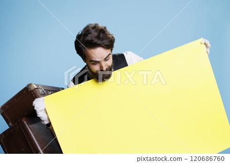 Hotel worker stands with empty poster in studio, holding blank banner with copyspace to create marketing ad campaign. Bellhop dressed in formal attire presents yellow banner on camera. Hotel worker stands with empty poster in studio, holding blank banner with copyspace to create marketing ad campaign. Bellhop dressed in formal attire presents yellow banner on camera. 120686760