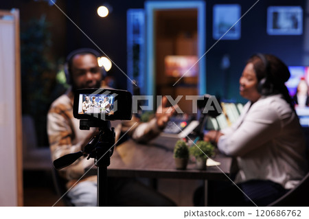 Black man creates vlogs with a digital camera and produces podcasts with professional audio gear. He interviews guests in his home studio, filming with a tripod. 120686762