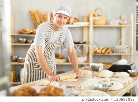 Portrait of young male baker wearing in apron working in bakehouse, rolling out dough in kitchen 120692052