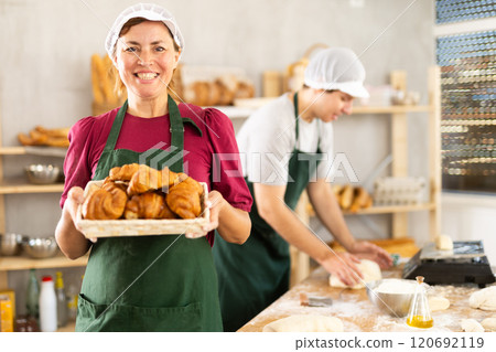 Portrait of happy female baker working in bakehouse, holding tray with fresh bakery goods on kitchen 120692119