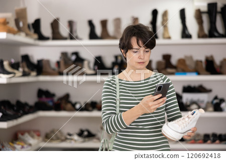 Female customer examines shoes in store and scans label with phone camera. 120692418