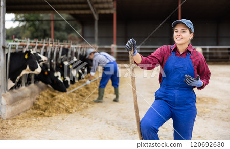 Portrait of smiling European female farmer in uniform with rake during work on dairy farm Portrait of smiling European female farmer in uniform with rake during work on dairy farm 120692680