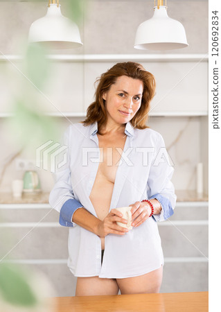 Topless woman in underpants and shirt stands in kitchen and covers bare chest with cloth of jacket. 120692834