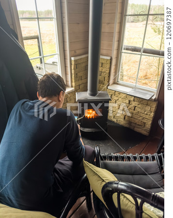 Man enjoying warmth by the wood stove in a cozy cabin with a scenic view. High quality photo 120697387