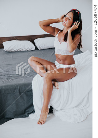 Tanned woman in lingerie listening to music in headphones on bed in bedroom 120697474