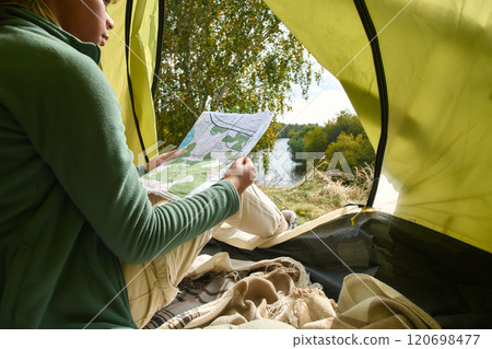 Medium close up of map in hands of unrecognizable female camper resting in cozy tent set on hill 120698477