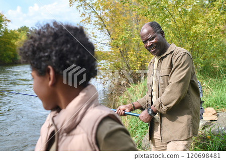 African American father with spinning in hands looking at his son while they fishing together 120698481
