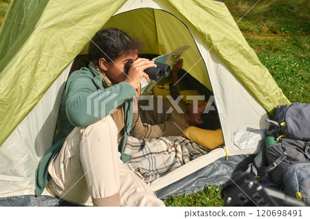 Mixed race teen girl sitting in tent and using binoculars while her little brother resting in tent and reading map 120698491
