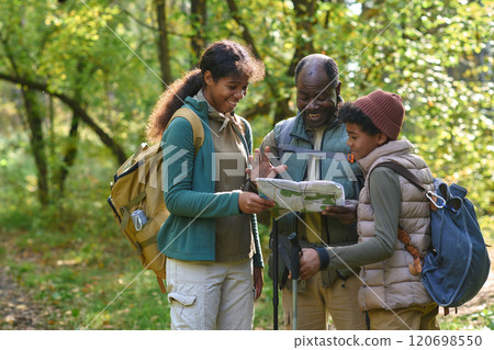 African American head of family choosing area for camp on map while his lovely and curious kids standing next to him and smiling African American head of family choosing area for camp on map while his lovely and curious kids standing next to him and smiling 120698550