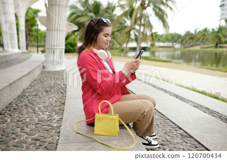 Young Woman Sitting Outside Checking Her Smartphone Young Woman Sitting Outside Checking Her Smartphone 120700434