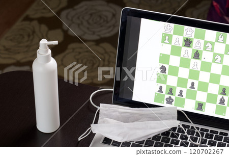 Chess task on laptop screen online. Near antibacterial spray and medical mask. Leisure in self-isolation. International chess day. 120702267