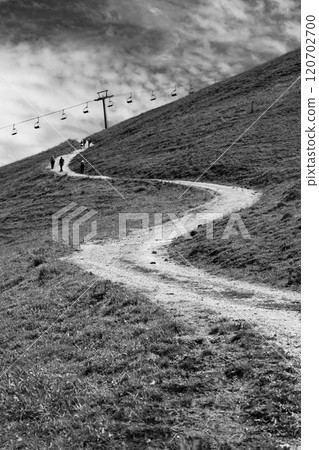 A winding alpine road stretches uphill, lined by grass and a cable car system above. A few hikers make their way along the path under a cloudy sky, enjoying the serene landscape. 120702700