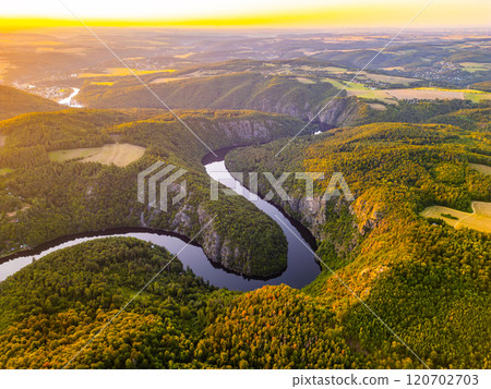 A breathtaking sunset bathes the Vltava River in golden hues, casting reflections on its winding path. Surrounded by lush hills and valleys, this serene moment captures the natural beauty of Czechia. 120702703