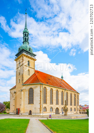 The Church of the Assumption of the Virgin Mary stands majestically in Most, showcasing late Gothic architecture under a bright blue sky and surrounded by lush greenery. 120702713