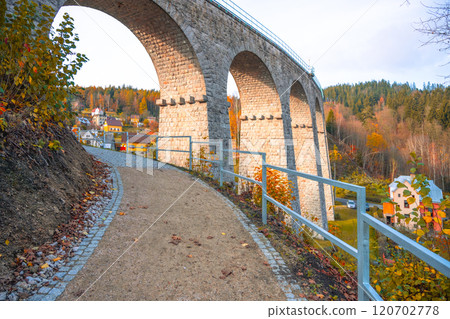 The railway viaduct in Smrzovka, nestled within the Jizera Mountains, stands proudly on a sunny autumn day, surrounded by colorful trees and charming village homes. 120702778