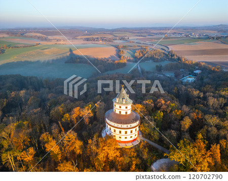 The Baroque Humprecht chateau stands elegantly above Sobotka, surrounded by vibrant autumn foliage as the sun rises, illuminating the landscape with a warm glow. 120702790