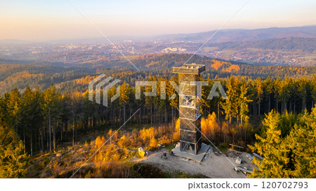 The wooden lookout tower stands tall against the vibrant autumn sunset near Liberec, surrounded by forested hills and distant views of the landscape bathed in warm colors. The wooden lookout tower stands tall against the vibrant autumn sunset near Liberec, surrounded by forested hills and distant views of the landscape bathed in warm colors. 120702793