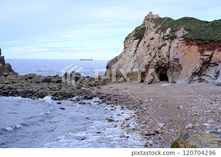 A rocky shoreline with a boat in the distance. The water is calm and the sky is overcast 120704296